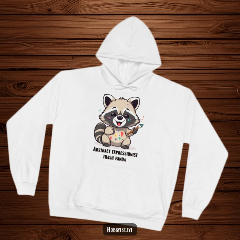 Funny hooded sweatshirt featuring a delighted raccoon artist with paint splatters, holding a brush, perfect for cozy creative sessions and art lovers.